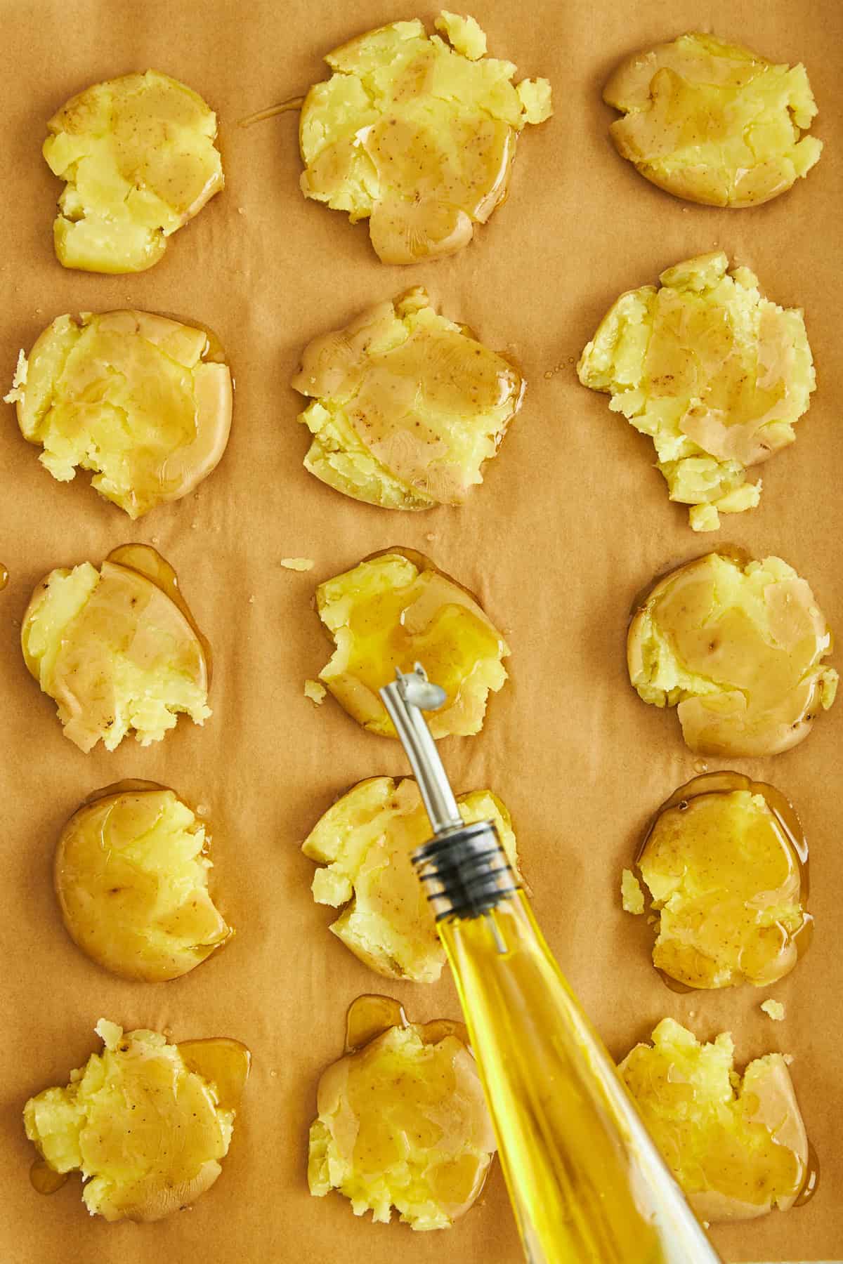 Olive oil being drizzled over smashed potatoes on a baking sheet. 