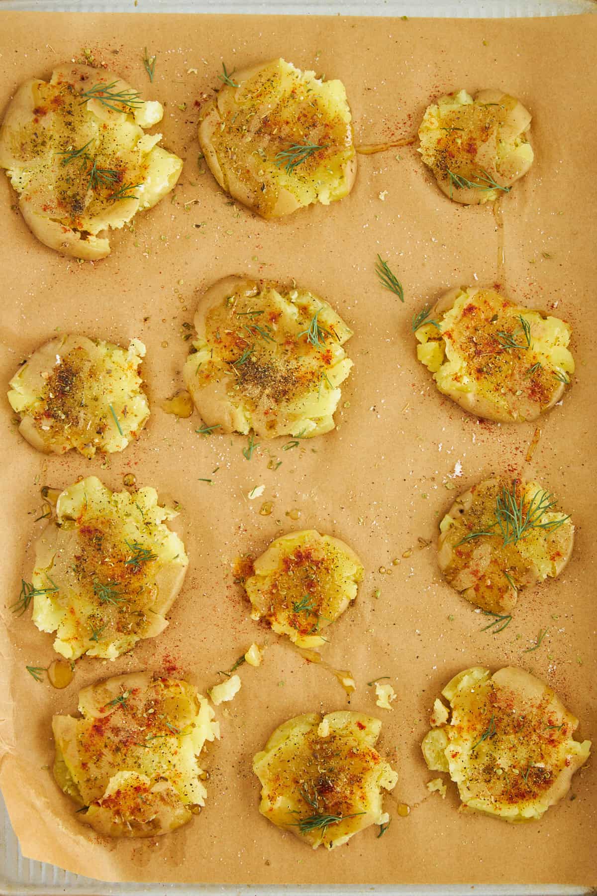 Uncooked smashed potatoes topped with oil and seasonings on a baking sheet. 