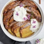 a white baking dish with baked cinnamon french toast topped with powdered sugar and marscapone cheese