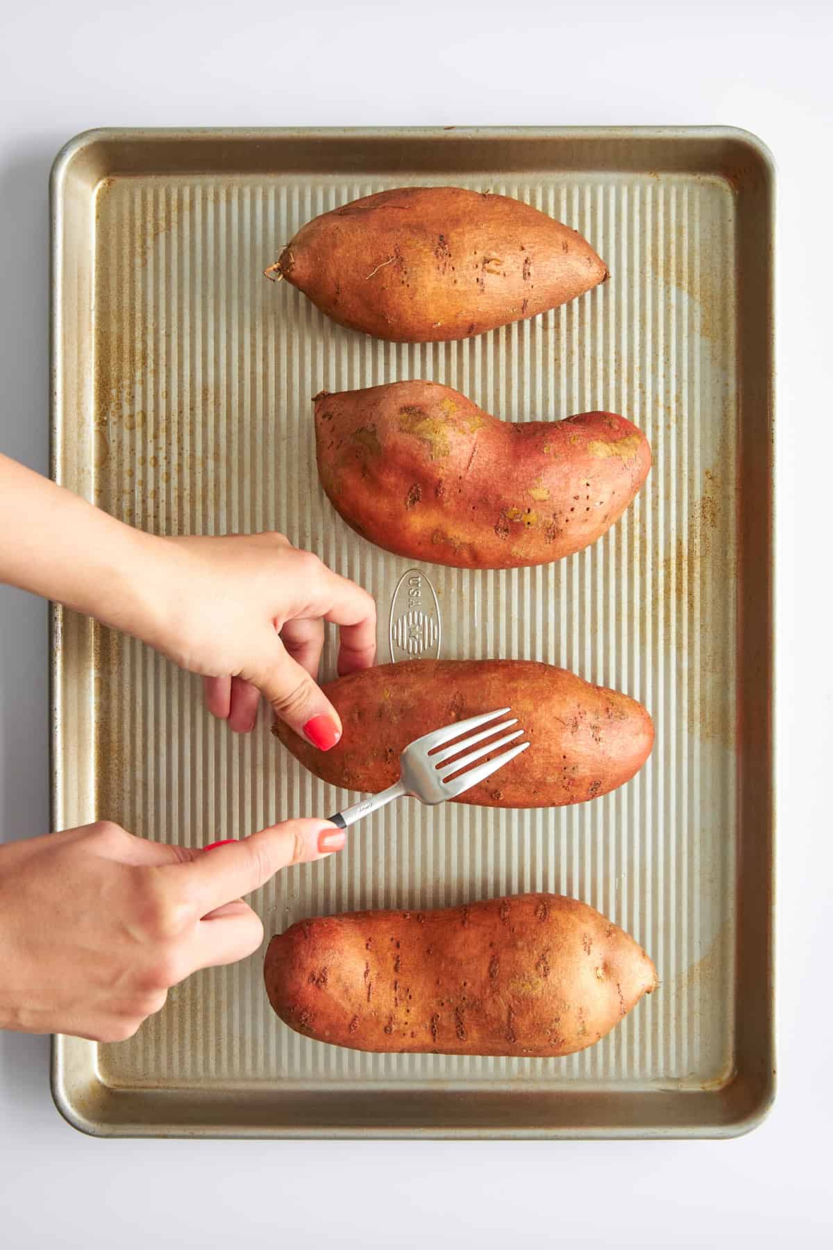 Four sweet potatoes on a baking sheet being pricked with forks. 