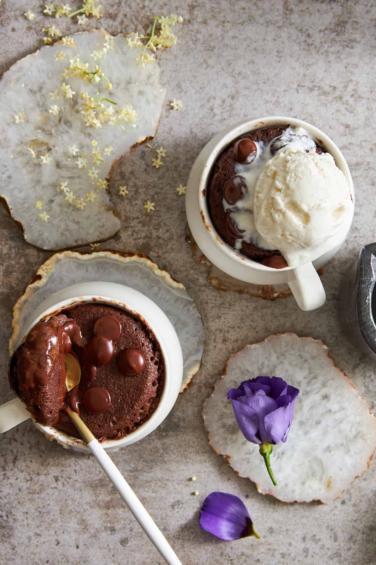 Two chocolate mug cakes, one with a spoon lifting a bite and the other topped with ice cream. 