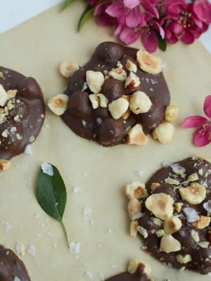 mounds of hazelnut chocolate crock pot candy topped with chopped hazelnuts and sea salt on a piece of parchment paper with pink flowers in the background