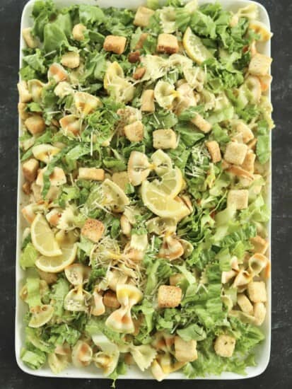 a rectangular white serving platter full with pesto pasta salad made with lettuce, noodles, Parmesan, croutons, and a pesto sauce topped with lemon wedges