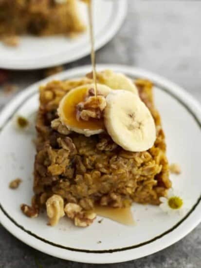 a piece of baked pumpkin oatmeal on a white plate topped with walnuts and banana coins with a drizzle of maple syrup being poured on top