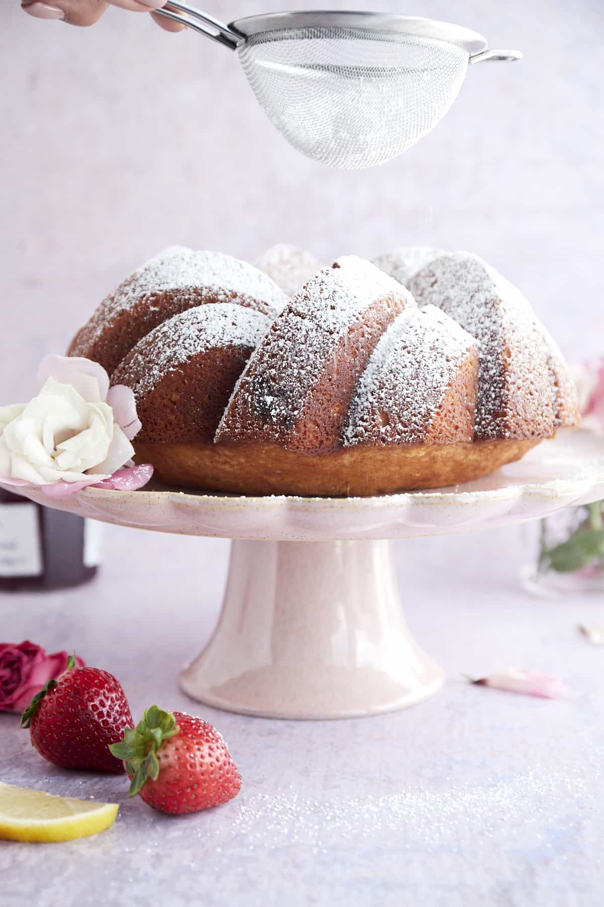 a strawberry lemon bundt cake on a cake stand being dusted with powdered sugar