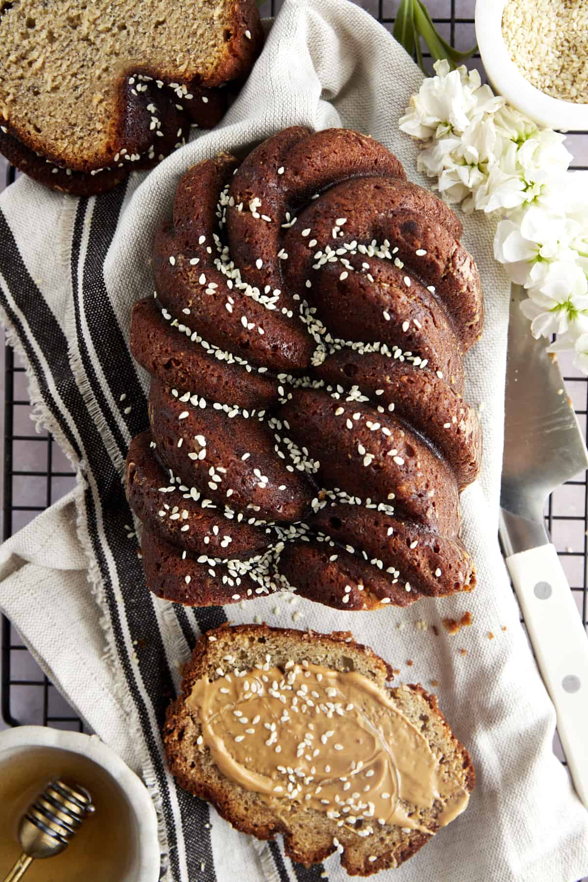 A loaf of tahini banana bread topped with honey and sesame seeds with a slice cut off and topped with nut butter and extra sesame seeds.