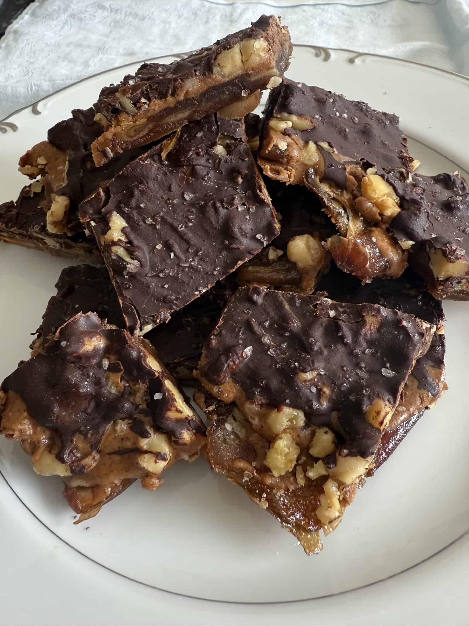 A plate of homemade toffee squares topped with melted chocolate and sprinkled with chopped nuts. The toffee is arranged in a stack, highlighting its rich texture and sugary gloss.