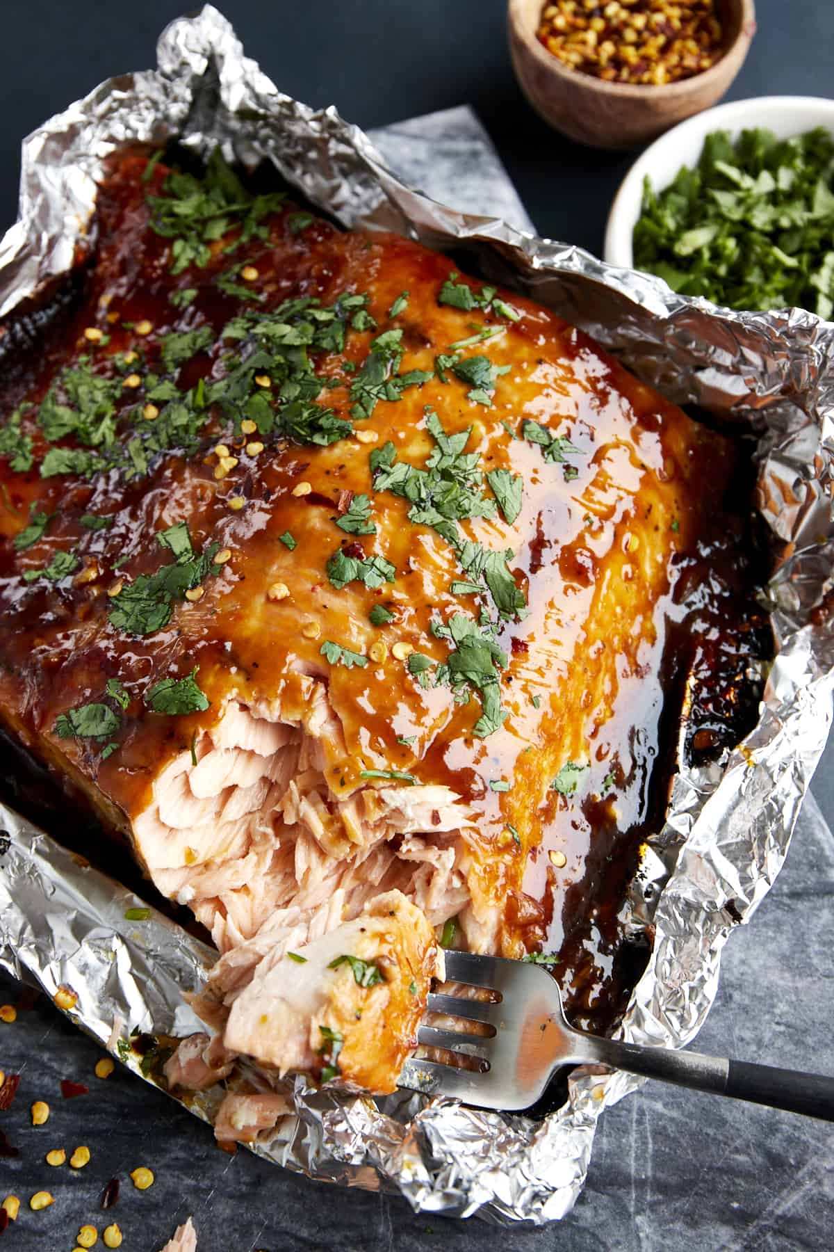 A fork tearing a bite of honey garlic salmon from a filet in a foil packet.