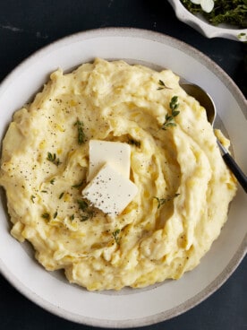 A bowl of the best mashed potatoes topped with butter, pepper, and herbs.