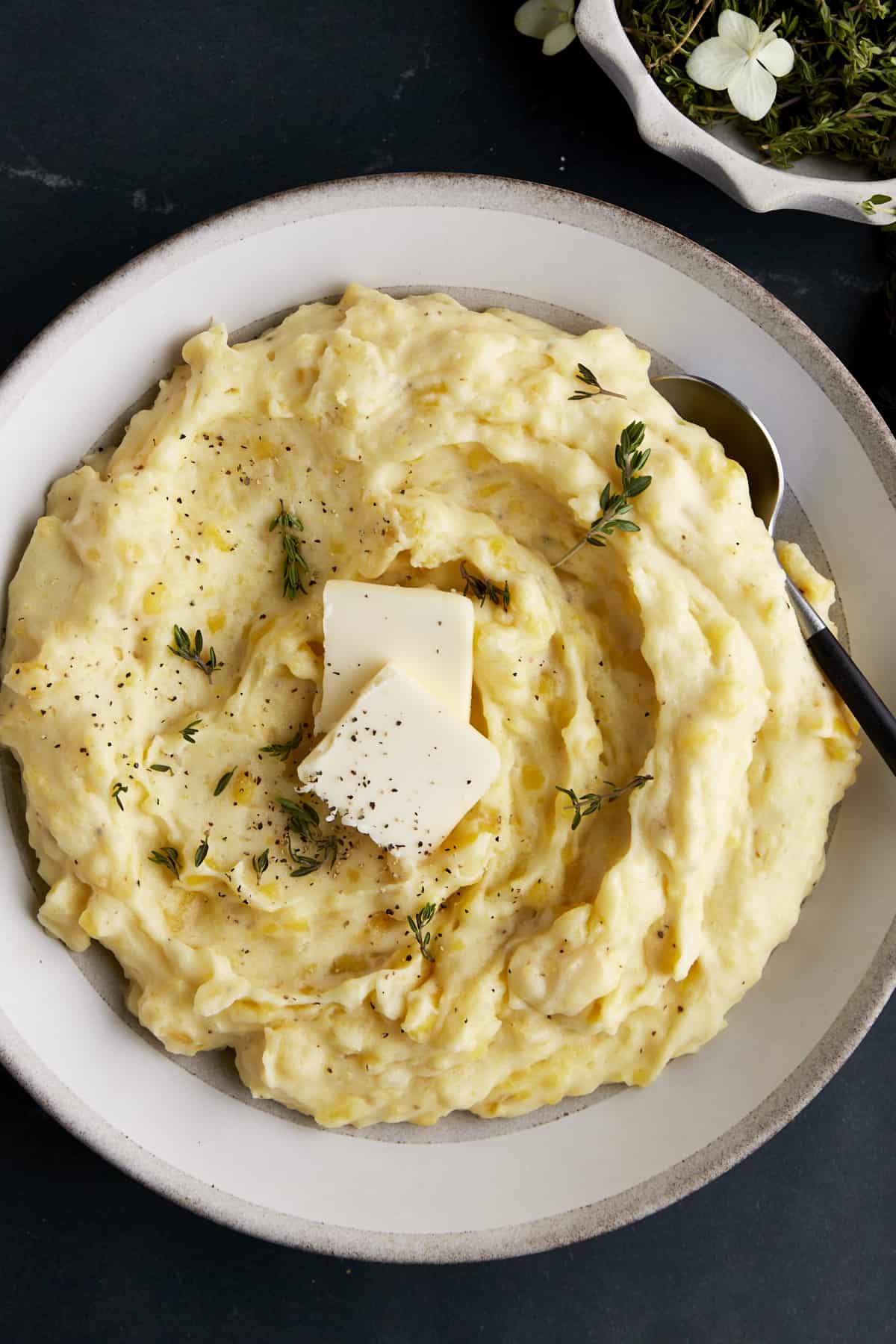 A bowl of the best mashed potatoes topped with butter, pepper, and herbs.