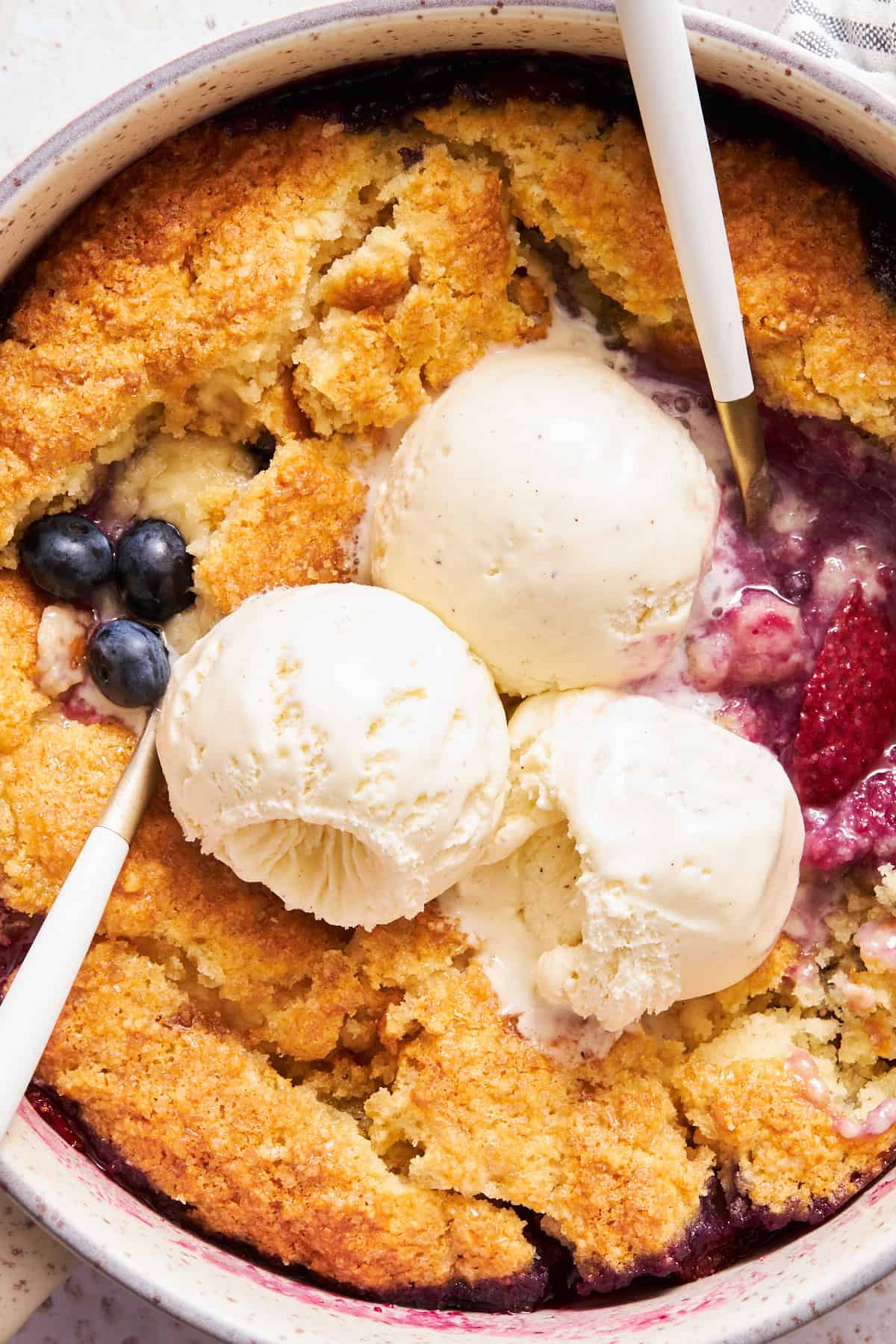 Close up view of two spoons in a mixed berry cobbler topped with three scoops of ice cream. 