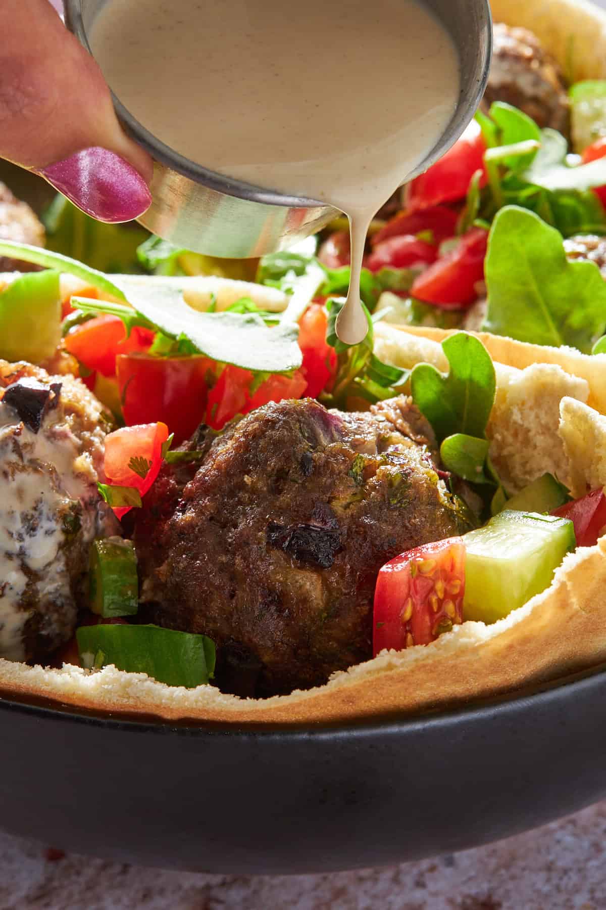 Tahini sauce being poured over a pita stuffed with Mediterranean beef meatballs and salad. 