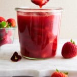 A spoon lifting a scoop of strawberry sauce from a jar.