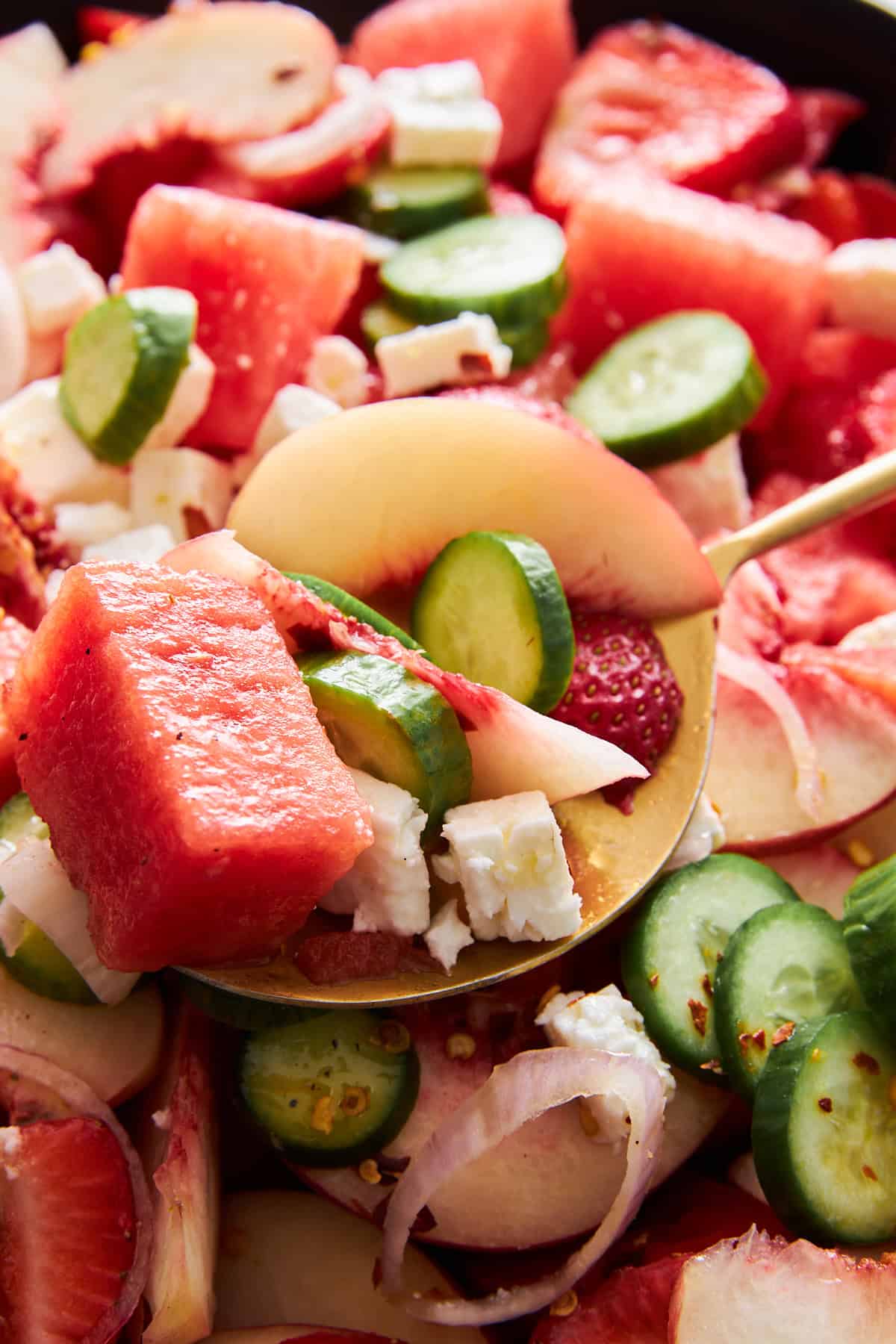 A spoon lifting a scoop of watermelon salad with feta, cucumbers, strawberries, and peaches. 