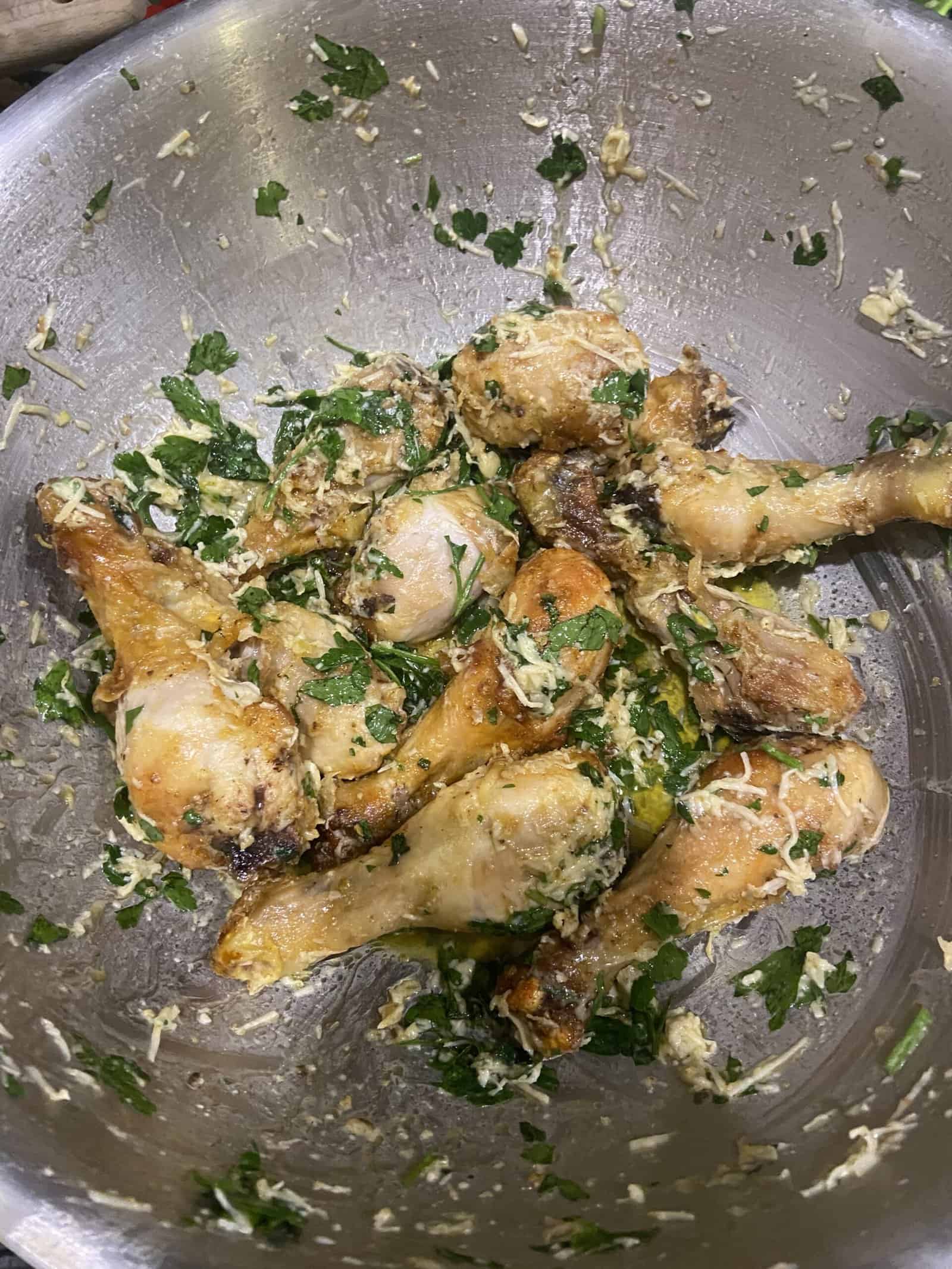 Cooked chicken drumsticks tossed with grated cheese and fresh chopped herbs in a stainless steel bowl.