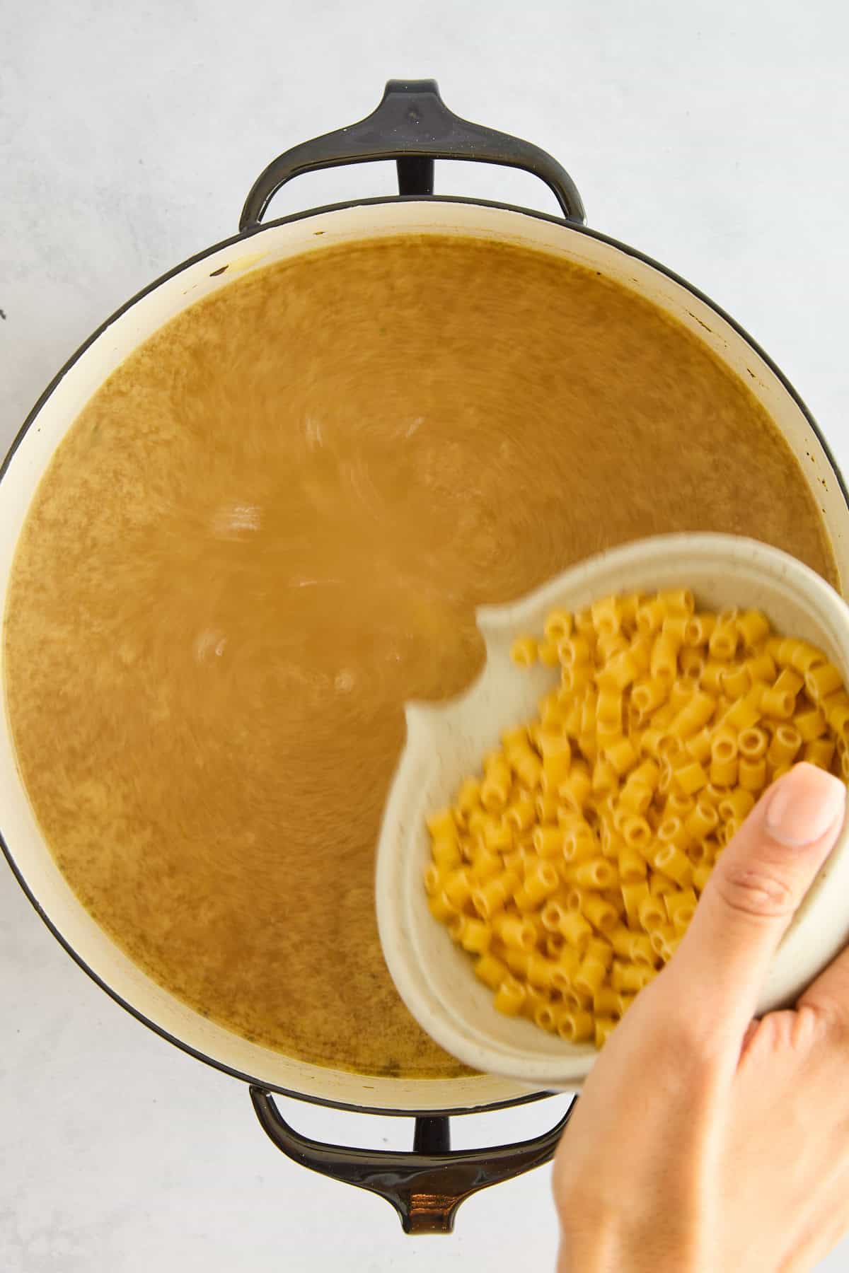 Raw ditalini pasta being poured into a pot of chicken noodle soup. 