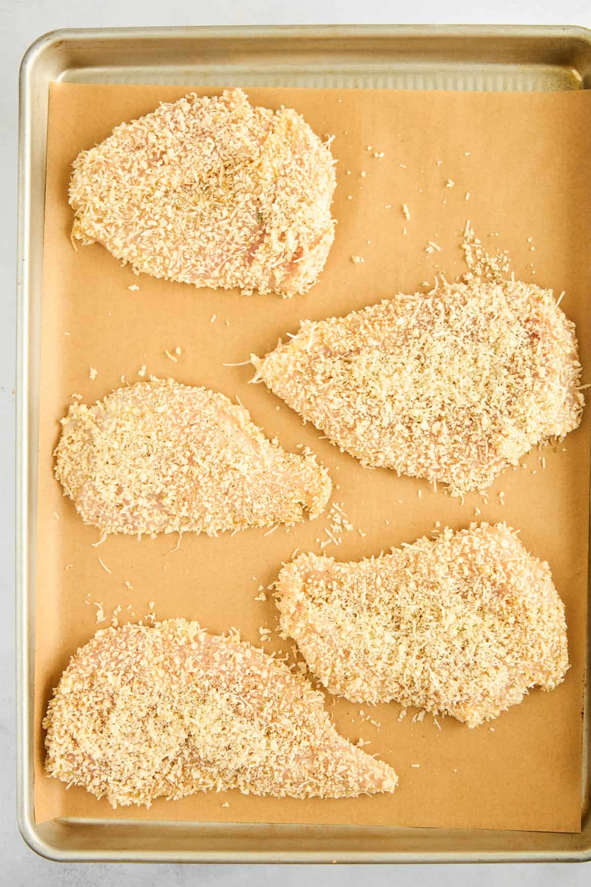 Five raw breaded chicken pieces on a lined baking sheet. 