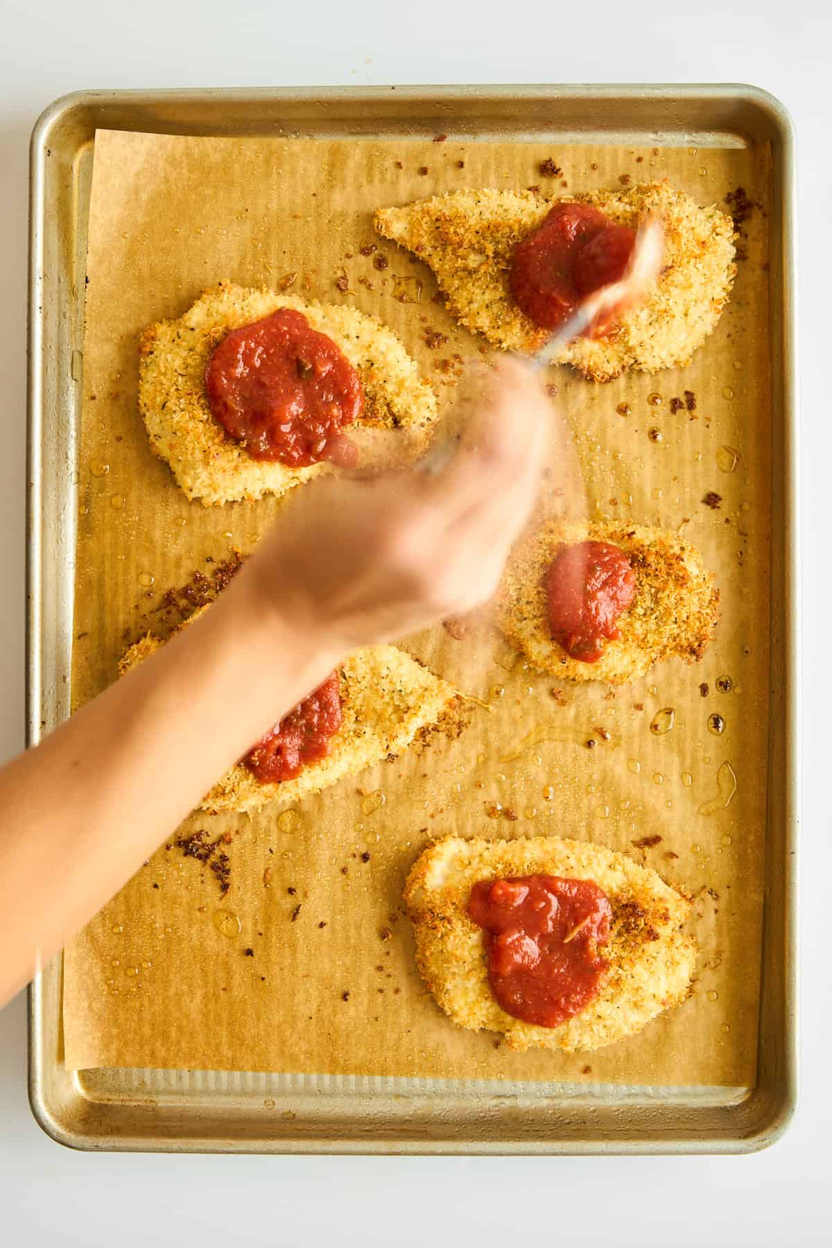 Marinara being spooned over breaded chicken breasts on a baking sheet. 