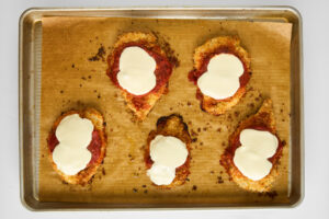 Five pieces of baked chicken parmesan on a baking sheet.