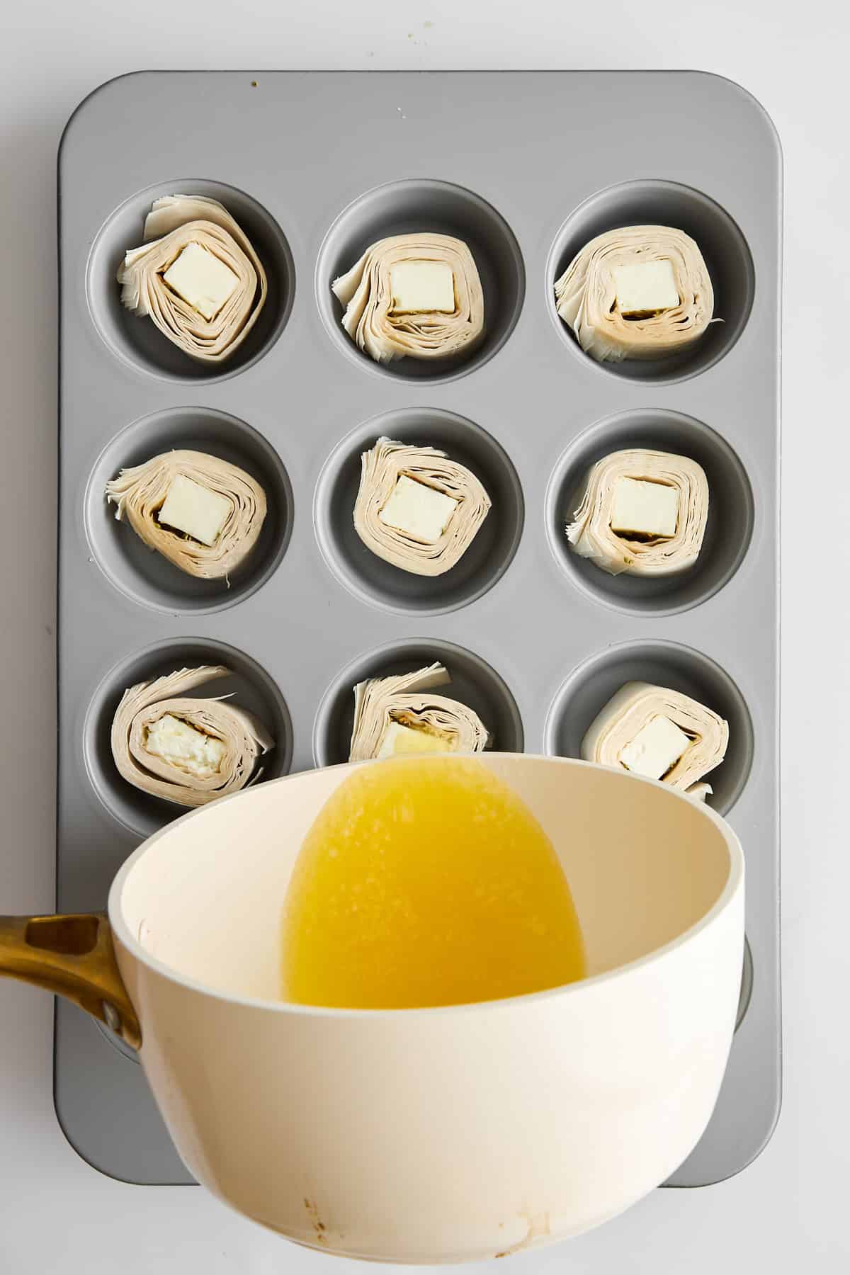 Feta phyllo rolls in a muffin pan being topped with butter. 