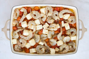 Raw shrimp and feta cubes in a baking dish over baked tomatoes.