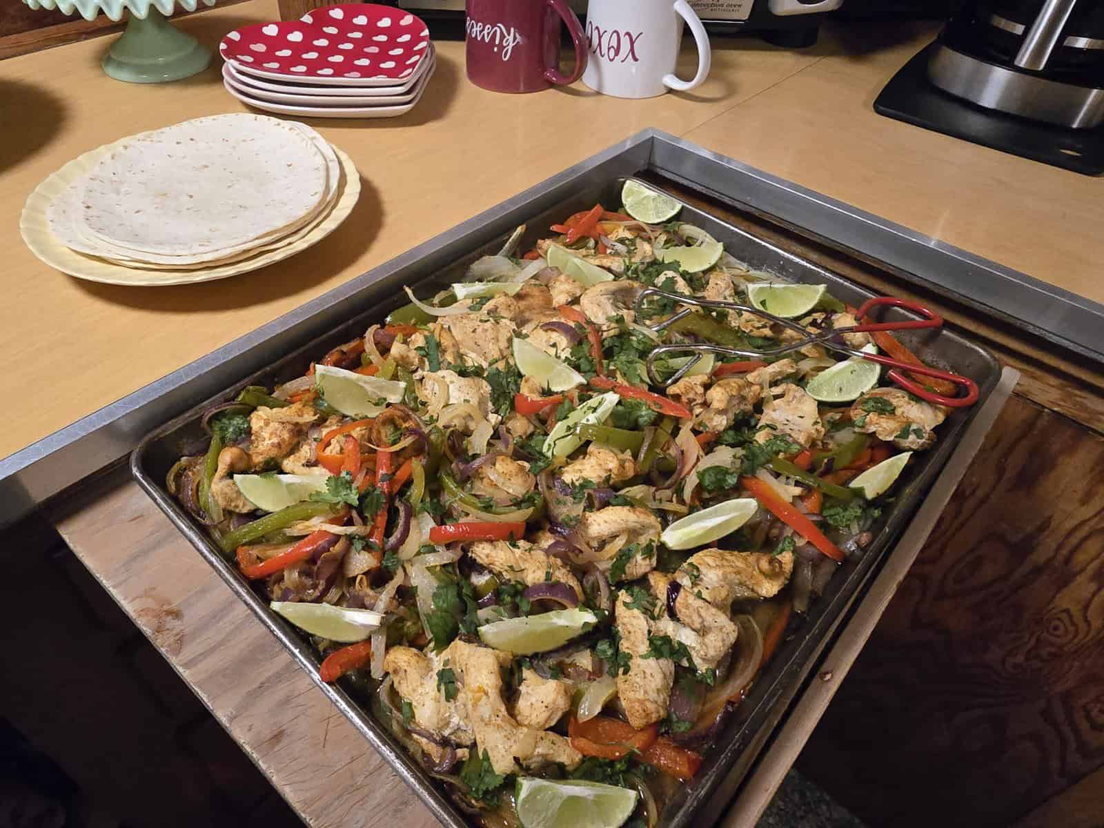 A tray of chicken fajitas with sliced lime wedges, colorful bell peppers, and cilantro is on a countertop. Nearby are stacked tortillas and mugs with decorative patterns.