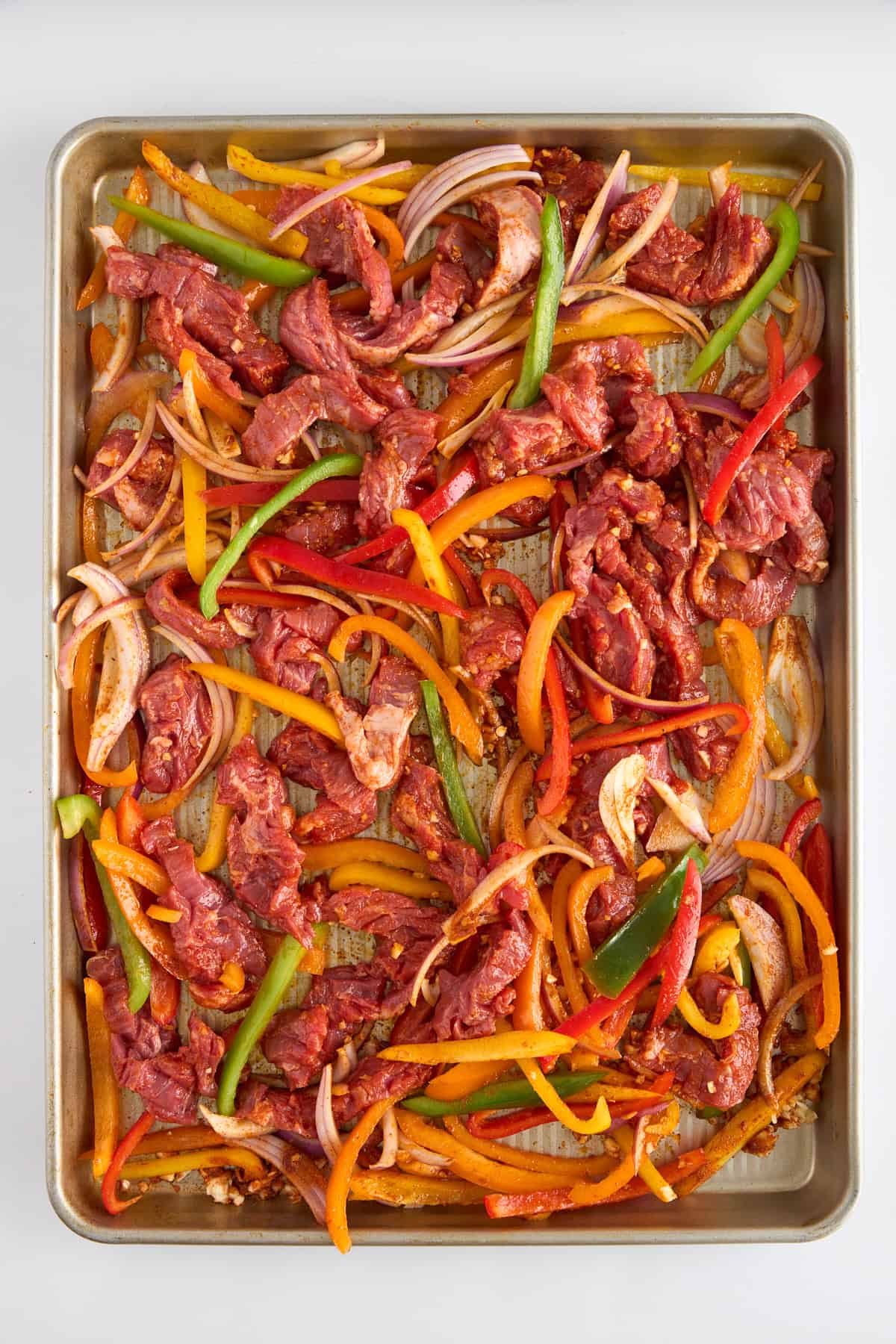 Raw fajita coated steak strips, bell peppers, and onions on a baking sheet. 