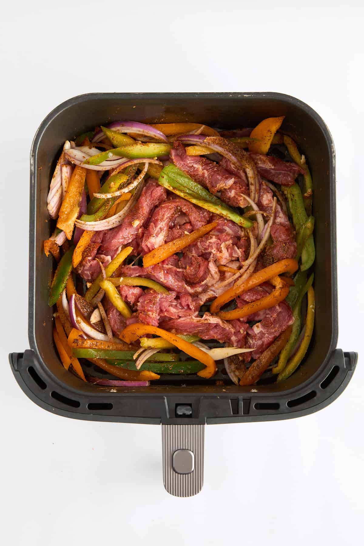 Raw steak strips, bell peppers, and onions in an air fryer basket. 