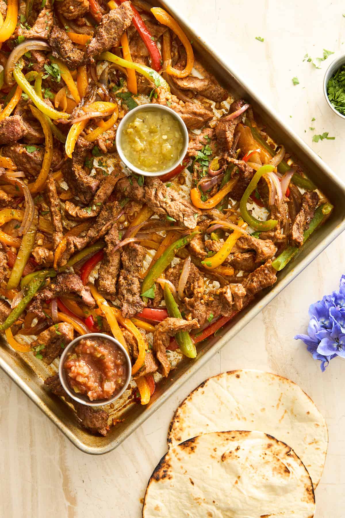 A baking sheet full of fajita steak, peppers, and onions with a cup of salsa verde and a cup of red salsa. 