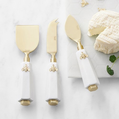 Three gold and white cheese knives with bee decorations.