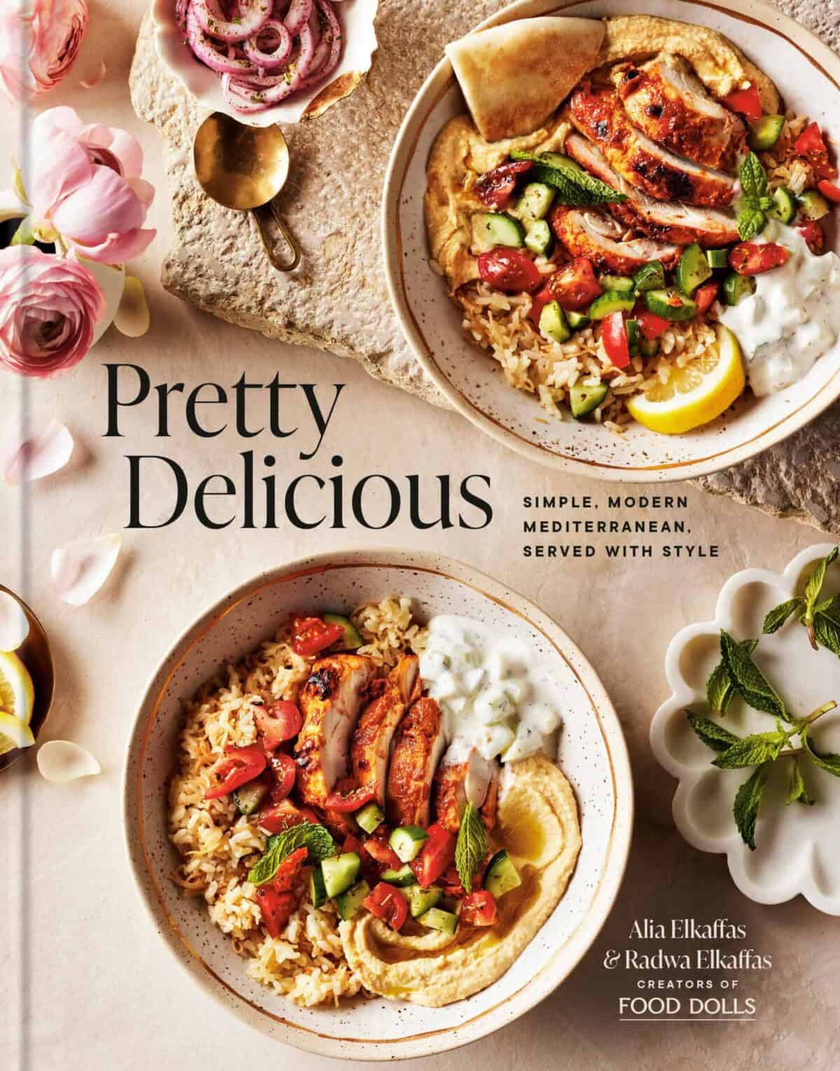 A cookbook titled Pretty Delicious featuring Mediterranean dishes. The cover shows two bowls with rice, grilled vegetables, falafel, and pita, garnished with lemon and herbs. Pink flowers and a small dish with yogurt are nearby.