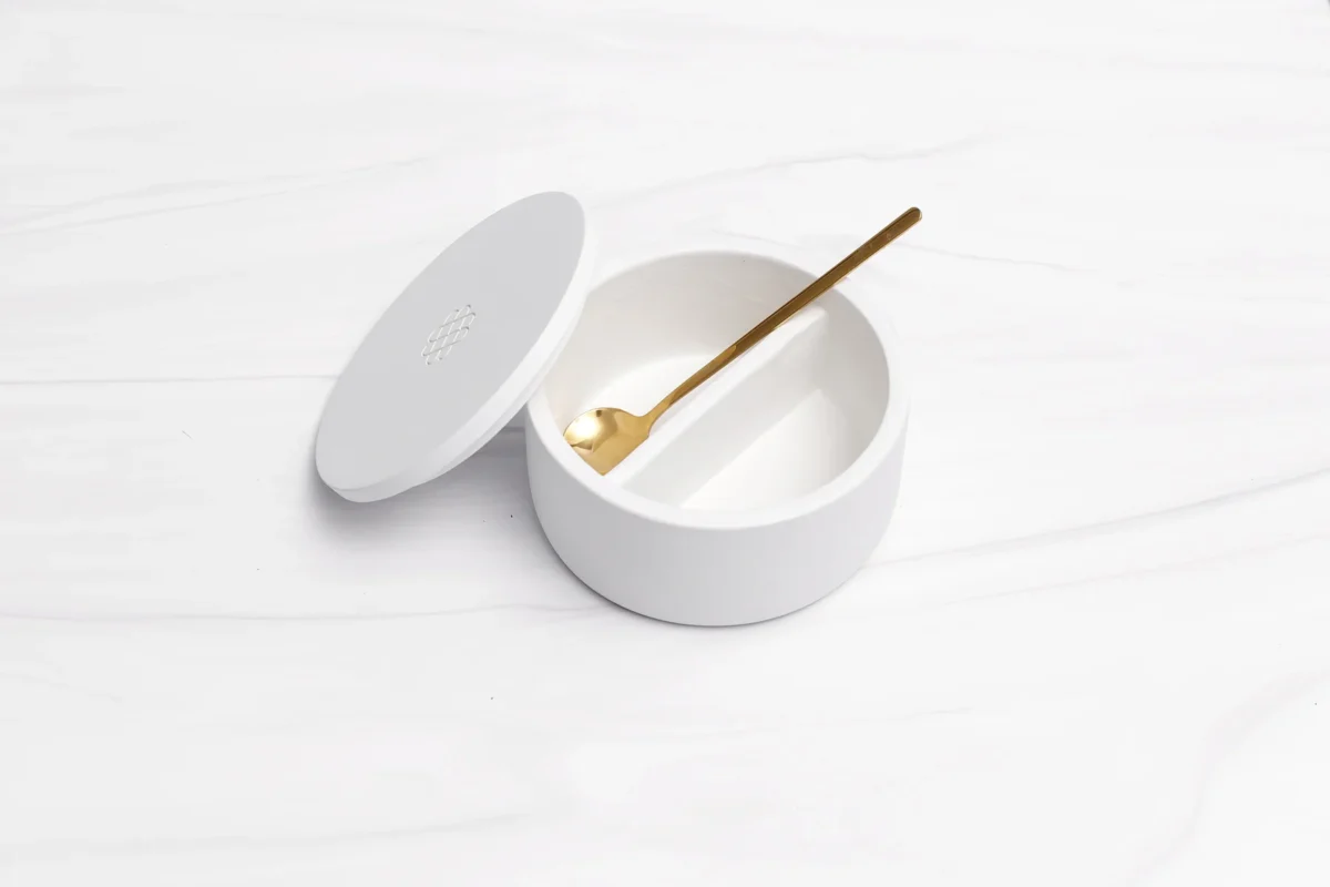 A white, round porcelain bowl with a lid sits open on a white marble surface. 